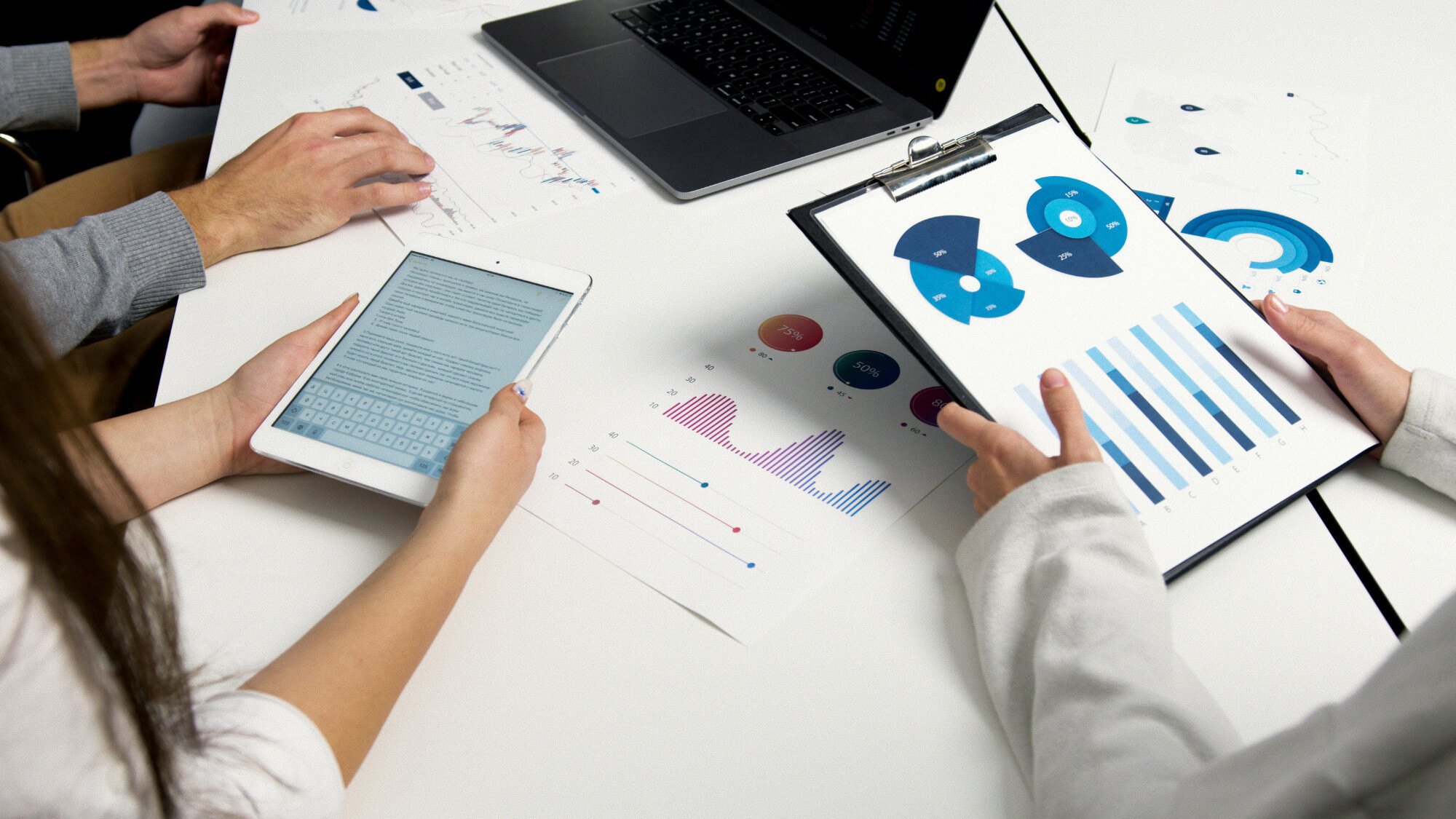 People sitting at desk in front of laptop holding tablets and paper stat charts.