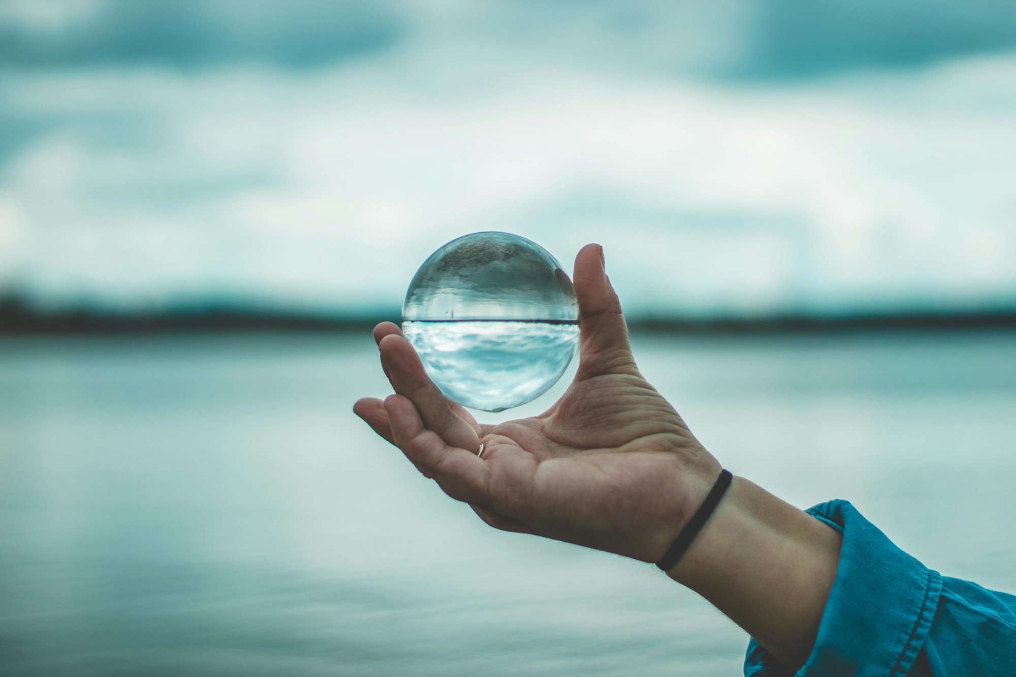 Hand holding circular glass ball in front of water horizon.