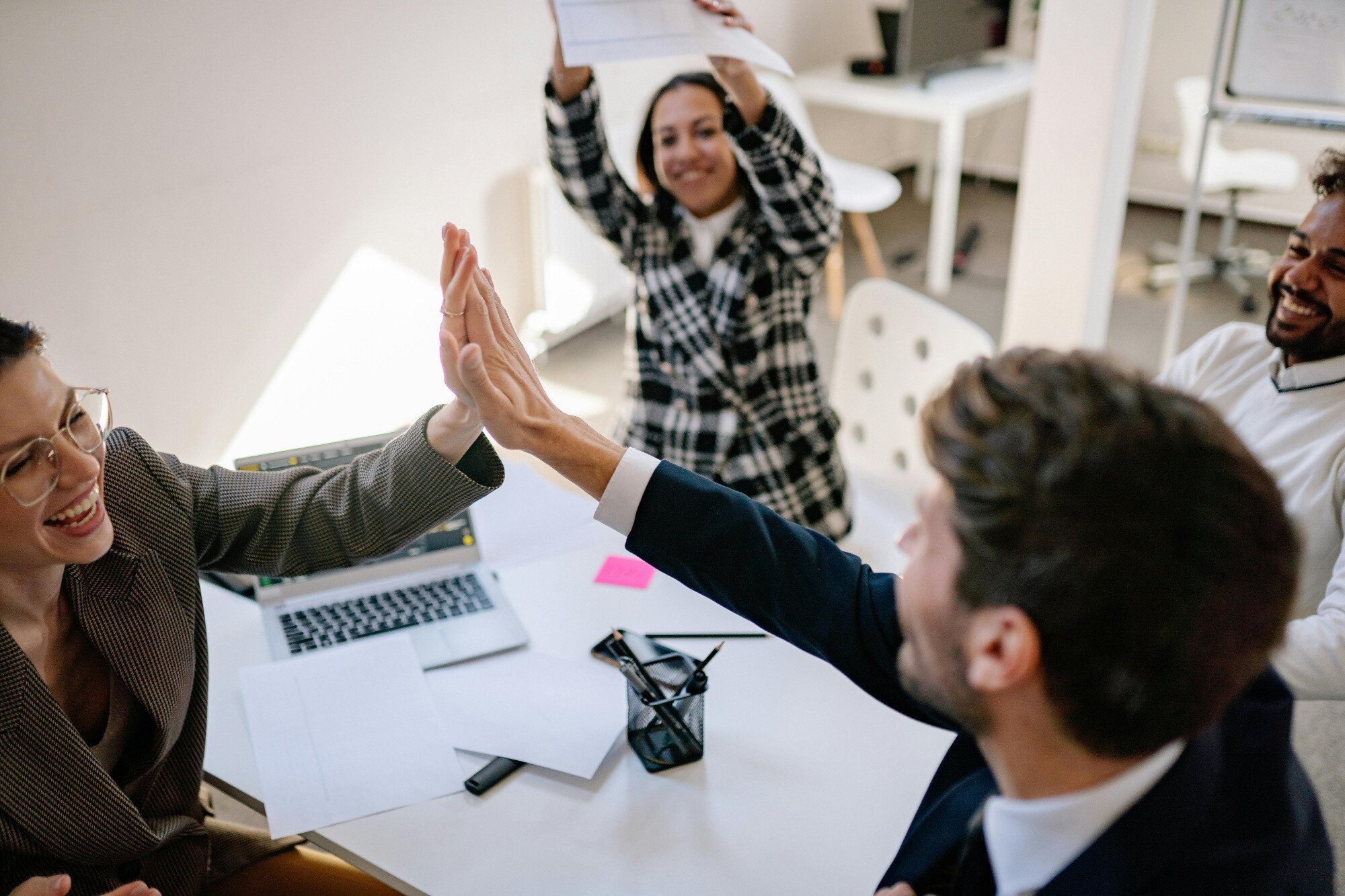 Two office workers sitting at desk high-fiving with another celebrating in the background.