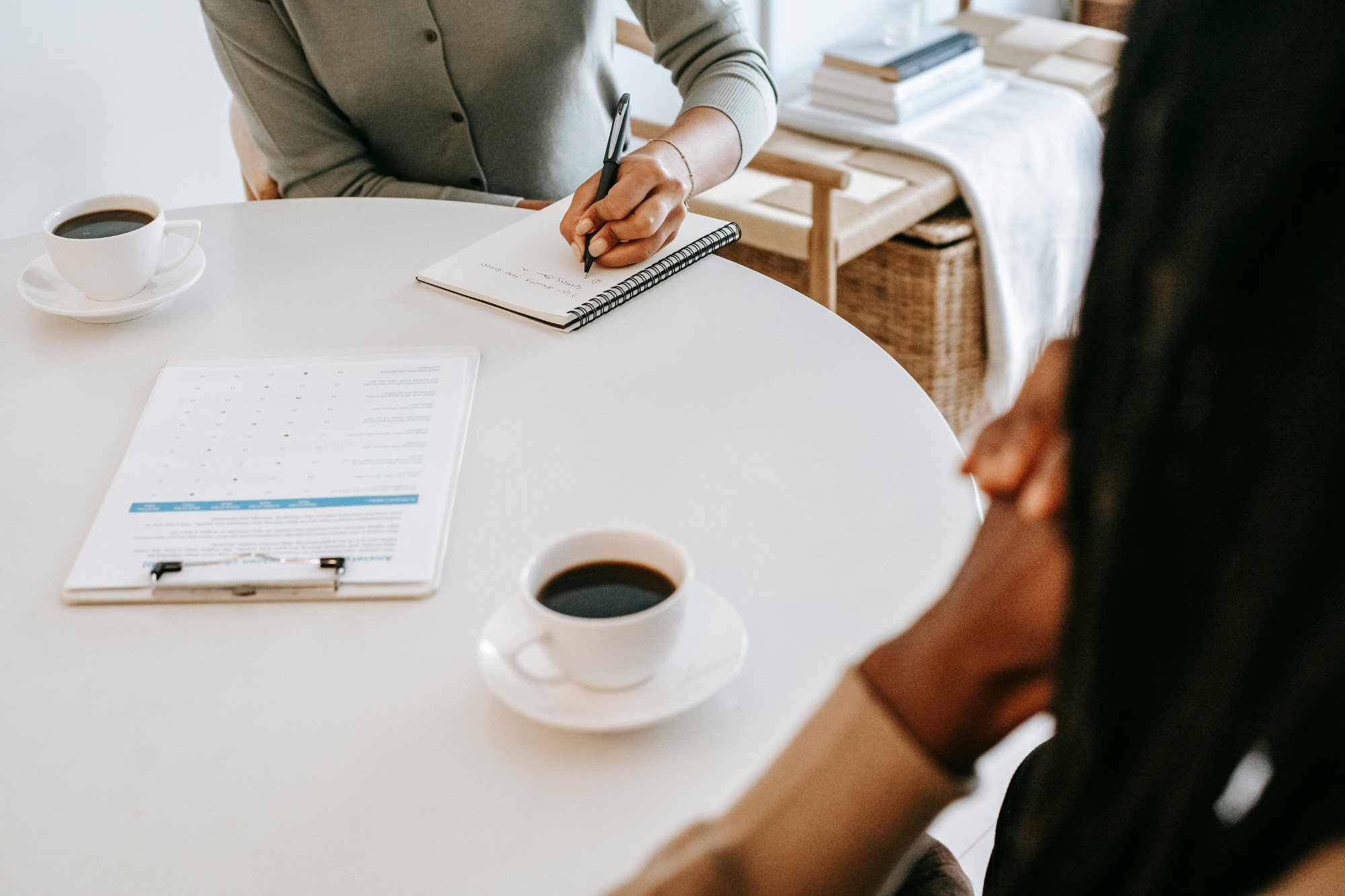 Two office workers sitting at round table with coffee, while one writes on a notepad.