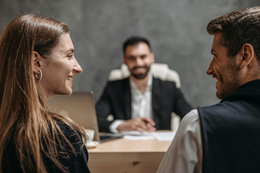 Two office workers smiling at each other while other office worker sits across the desk from them.